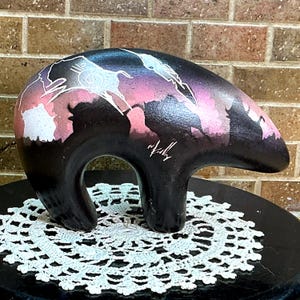 May include: A smooth, black, bear-shaped sculpture featuring a painted scene of silhouettes of bison against a pink and purple sunset sky. The artwork includes white line drawings of animals and a signature in white ink on the side of the bear.