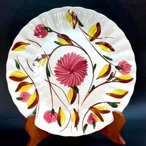 May include: White ceramic plate with a ruffled edge, featuring a hand-painted floral design. The design includes a large pink flower, smaller pink roses, and yellow and brown leaves on brown stems. The plate is displayed on a wooden stand.