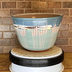 May include: A ceramic bowl featuring a hand-painted landscape design with light blue sky, green fields, and pale pink cattails. The bowl has a wide, open top and a tapered base, showcasing a smooth, glazed finish with visible brushwork details.