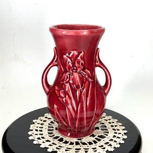 May include: A burgundy-colored ceramic vase with a raised iris flower design. The vase has two handles and a flared rim. The vase is approximately 8 inches tall.