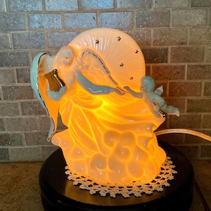May include: A white ceramic illuminated angel figurine featuring a large angel holding a golden lyre and a smaller cherub reaching out. The piece has a circular backdrop with gold star cutouts and emits a warm orange glow from within.