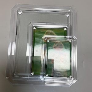 May include: Three clear acrylic picture frames of varying sizes. Each frame has a rectangular opening with a beveled edge and a decorative geometric design at each corner. The frames are stacked, with a photo visible in each.