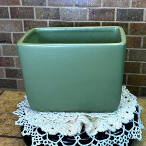 May include: A rectangular sage green ceramic planter with a smooth, matte finish and rounded corners. The container features a wide, open top and solid walls, providing a clean and simple structure for holding various items or small plants.