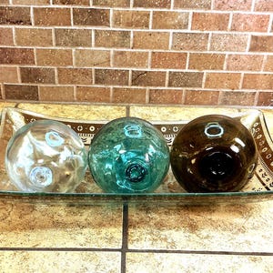 May include: A rectangular glass tray with a brown patterned border holds three spherical glass floats. The floats are arranged in a row, featuring a clear glass sphere, a translucent teal glass sphere, and a dark brown glass sphere.