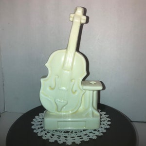 May include: A white ceramic figurine of a violin with a stand. The violin has a detailed design and the stand has a small hole for holding pencils or pens.