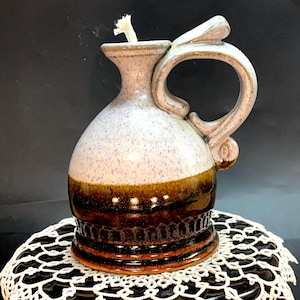 May include: A handmade ceramic oil lamp with a handle. The lamp features a light purple and brown color scheme with a white wick. The base has a decorative pattern. The lamp is designed for use with oil.