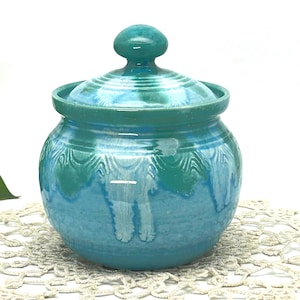 May include: A ceramic jar with a lid, featuring a turquoise and teal color scheme. The jar has a rounded shape with a lid and a small knob. The glaze has a textured, dripped effect, creating a unique, handcrafted appearance. The jar is likely used for storage.