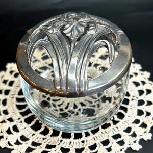 May include: A clear glass jar with a decorative silver lid. The lid features a floral design with long, slender leaves and a central flower. The jar sits on a white lace doily against a dark background.