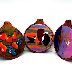 May include: Set of three round wooden serving boards with colorful painted designs. One board features red apples, orange pears, and blue grapes. Another has a still life with glasses and a pitcher. The third board has a purple and gold design.