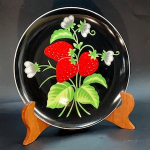 May include: A black ceramic plate with a white rim, featuring a vibrant design of red strawberries, green leaves, and white flowers. The plate is displayed on a wooden stand, showcasing its decorative fruit motif.