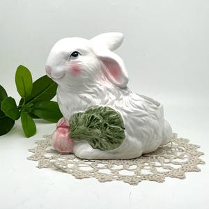 May include: A white ceramic bunny planter with pink accents on the ears and cheeks, holding a green lettuce. The bunny is sitting on a beige lace doily. The planter is a decorative item.
