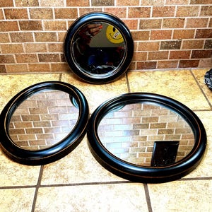 May include: Three mirrors with black circular and oval frames featuring thin gold-toned pinstripe detailing. The frames have a smooth finish and are arranged in a group. The reflective glass surfaces show a clear view of the surrounding environment.