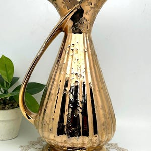 May include: A gold ceramic vase with a textured surface and a curved handle. The vase has a wide mouth and a narrow base.