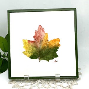 May include: Square ceramic tile with a watercolor painting of a fall leaf. The leaf transitions from red to yellow to green. The tile has a dark green border and is displayed on a decorative stand.
