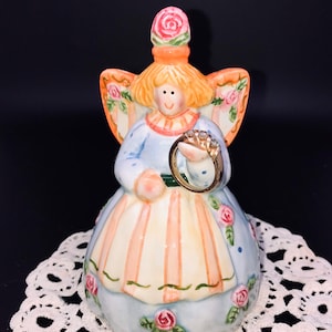 May include: A ceramic angel figurine with a blue dress, pink and orange accents, and a floral design. The angel is holding a ring in her hand.