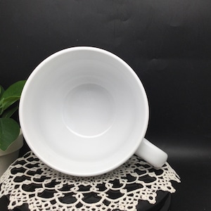 Puede incluir: Taza de cer&aacute;mica blanca con una gran forma de cuenco y un asa. La taza est&aacute; vac&iacute;a y se encuentra sobre una puntilla blanca.