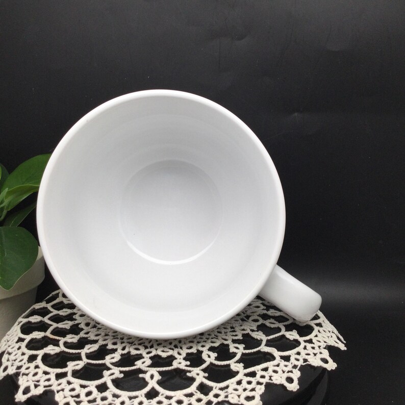 Puede incluir: Taza de cer&aacute;mica blanca con una gran forma de cuenco y un asa. La taza est&aacute; vac&iacute;a y se encuentra sobre una puntilla blanca.