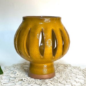 May include: A handcrafted, yellow ceramic lantern with a textured surface. The lantern features vertical cut-out openings and a pedestal base with a brown rim. The interior is visible through the openings, creating a play of light and shadow.