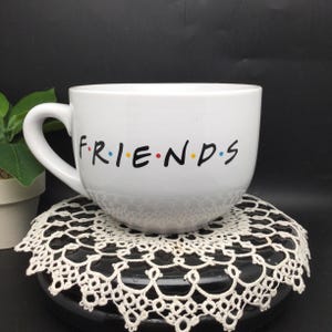 Puede incluir: Taza de cer&aacute;mica blanca con un logotipo "FRIENDS" negro y puntos de colores. La taza est&aacute; sobre un encaje blanco.