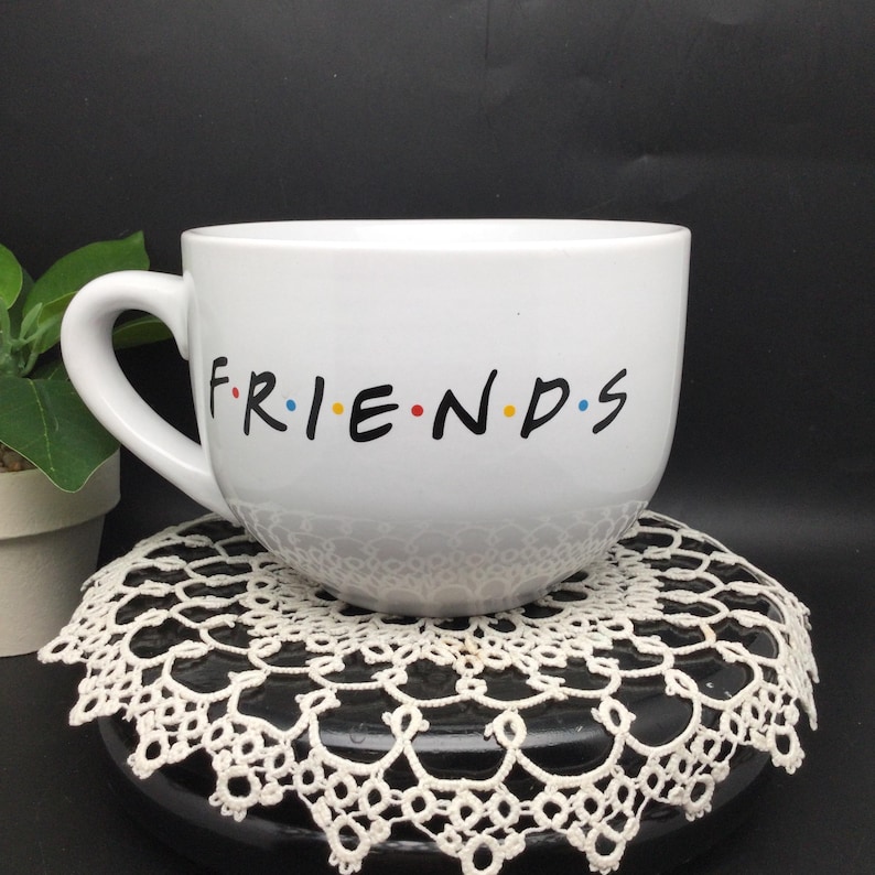 Puede incluir: Taza de cer&aacute;mica blanca con un logotipo "FRIENDS" negro y puntos de colores. La taza est&aacute; sobre un encaje blanco.