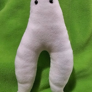 Könnte beinhalten: Weißes Plüschtier in Form einer Person mit großen Beinen und schwarzen Augen. Das Spielzeug sitzt auf einer grünen Oberfläche.