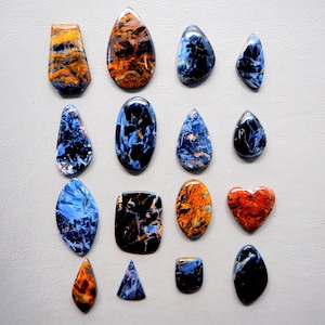 Puede incluir: Una colección de piedras preciosas pulidas en varias formas, incluyendo lágrimas, óvalos y un corazón. Las piedras muestran una mezcla de colores, con azules, naranjas y marrones prominentes. Las piedras preciosas están dispuestas sobre una superficie gris claro.