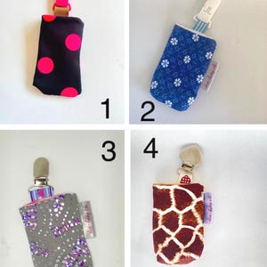 May include: Four baby pacifier holders with different patterns. One is black with pink polka dots, another is blue with white flowers, a third is gray with purple accents, and the last is brown with a giraffe print. Each has a clip.