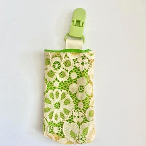 May include: A small, rectangular holder with a lime green interior and a cream-colored floral lace exterior. It has a lime green clip and a cream-colored ribbon. The holder is designed to hold small items.