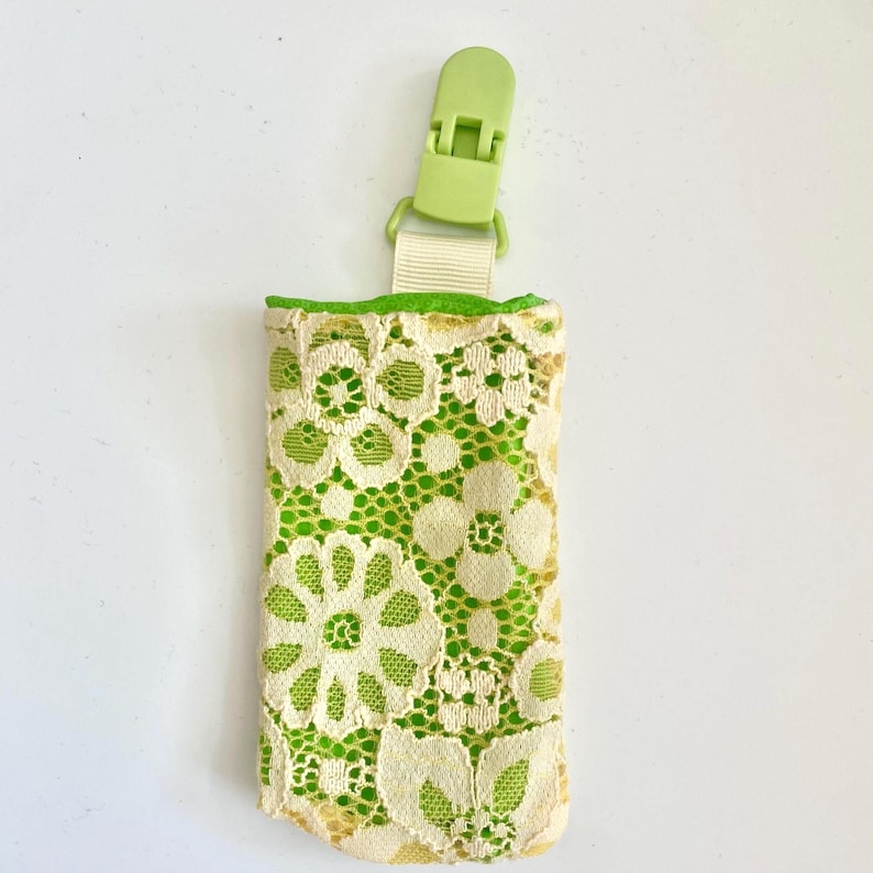 May include: A small, rectangular holder with a lime green interior and a cream-colored floral lace exterior. It has a lime green clip and a cream-colored ribbon. The holder is designed to hold small items.