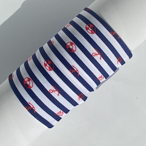 Può includere: Un braccialetto in tessuto con un motivo a righe orizzontali blu navy e bianche. Il design è arricchito da piccole icone nautiche rosse ripetute, tra cui ancore, salvagenti e barche a vela, distribuite sulla superficie a righe.