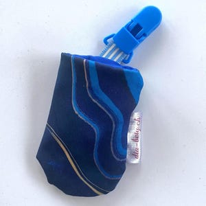 Puede incluir: Pequeña bolsa de tela azul marino y azul con un clip de plástico azul. La bolsa tiene un patrón veteado en tonos azules y dorados. Se adjunta una pequeña etiqueta blanca con texto rojo. El clip está sujeto a una tela a rayas.
