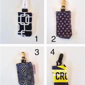 May include: Four pacifier clips with different fabric patterns. The clips have a plastic or metal clip at the top and a fabric pouch at the bottom. The patterns include a black and white geometric design, a navy pattern, a white polka dot pattern, and a yellow and black text design.