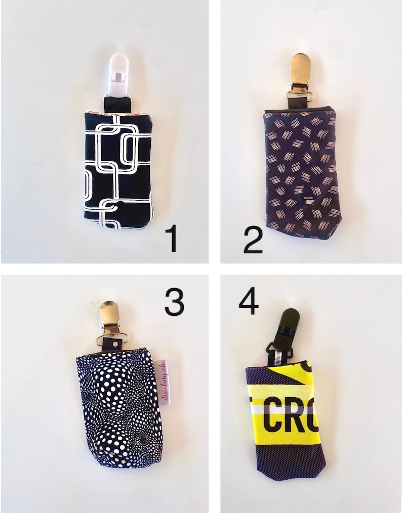 May include: Four pacifier clips with different fabric patterns. The clips have a plastic or metal clip at the top and a fabric pouch at the bottom. The patterns include a black and white geometric design, a navy pattern, a white polka dot pattern, and a yellow and black text design.