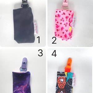 May include: Four different pacifier clips are displayed. The first is black, the second is pink with a floral pattern, the third is purple with a galaxy design, and the fourth is black with bear graphics. Each clip has a matching plastic clip.