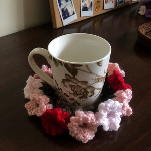 May include: A crocheted flower coaster in shades of pink, red and white, with a white mug with a floral pattern sitting on top.