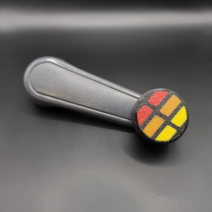 May include: Grey plastic window winder with a round, black knob. The knob has a colourful geometric design with red, orange, and yellow segments.