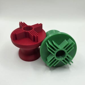 May include: Two 3D-printed objects. One is red, upright, with a cylindrical base and a top shaped like a cross with angled blades. The other is green, lying on its side, with a similar cross-shaped top. Both have a central hole.