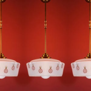 Könnte beinhalten: Drei Pendelleuchten mit weißen, schalenförmigen Schirmen. Jeder Schirm hat ein sich wiederholendes Muster aus roten und goldenen Ananas-Designs. Die Leuchten hängen an messingfarbenen Stangen vor einem roten Hintergrund.