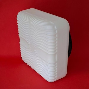 Può includere: Un apparecchio di illuminazione rettangolare bianco con un design a spirale e strutturato sulla parte anteriore. L'apparecchio ha angoli arrotondati e un retro nero, su sfondo rosso. Il design assomiglia a un'esplosione solare stilizzata.