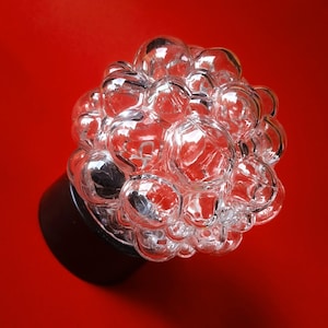 Pode incluir: Uma lâmpada decorativa de vidro transparente com um grupo de globos em forma de bolha. A lâmpada assenta numa base castanha escura sobre um fundo vermelho brilhante. As bolhas de vidro refletem a luz.