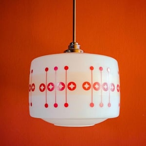 May include: A white pendant light with a cylindrical shade, decorated with red circles and diamond shapes. The light hangs from a brown rod. The background is a vibrant orange. A retro design.