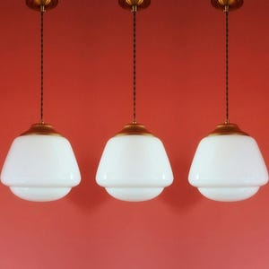 Könnte beinhalten: Drei Pendelleuchten mit weißen Glasschirmen und messingfarbener Hardware hängen vor einem roten Hintergrund. Jede Leuchte hat einen dekorativen Aufsatz und ein gedrehtes Kabel.