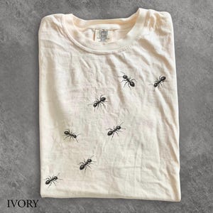 May include: An ivory-coloured crew neck t-shirt featuring a scattered pattern of small, black ant illustrations across the front. The inner neck label displays the text Comfort Colors 100% Cotton. The shirt has short sleeves and a standard fit.
