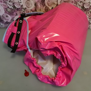 Puede incluir: Una cubierta de pañal de vinilo rosa con una correa negra y un borde elástico blanco. La cubierta está abierta y el interior es blanco.