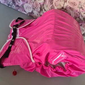 Puede incluir: Un pañal para adultos de color rosa con ribetes blancos y correas negras. El pañal tiene un acabado brillante y húmedo.