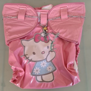 Puede incluir: Cubierta de pañal Hello Kitty rosa con una cadena y un candado unidos. La cubierta de pañal tiene un gráfico Hello Kitty blanco con un vestido azul y flores blancas.