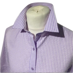Può includere: Una camicia abbottonata a quadri lilla e bianca. La camicia è esposta su un manichino, mettendo in risalto il colletto e i dettagli dei bottoni. Il tessuto ha una trama sottile e lo stile è classico e versatile.