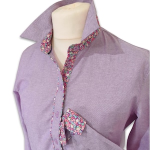 Könnte beinhalten: Ein helllila Hemd mit Knöpfen und einem floralen Muster am Kragen, den Manschetten und der Knopfleiste. Das Hemd hat einen klassischen Kragen und lange Ärmel. Der Blumendruck umfasst rosa, blaue und grüne Blumen auf dunklem Hintergrund.