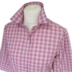 May include: A short-sleeved button-down shirt featuring a plaid pattern in shades of pink, white, and light blue. The shirt has a structured collar and a front placket with matching pink buttons, displayed on a mannequin torso.