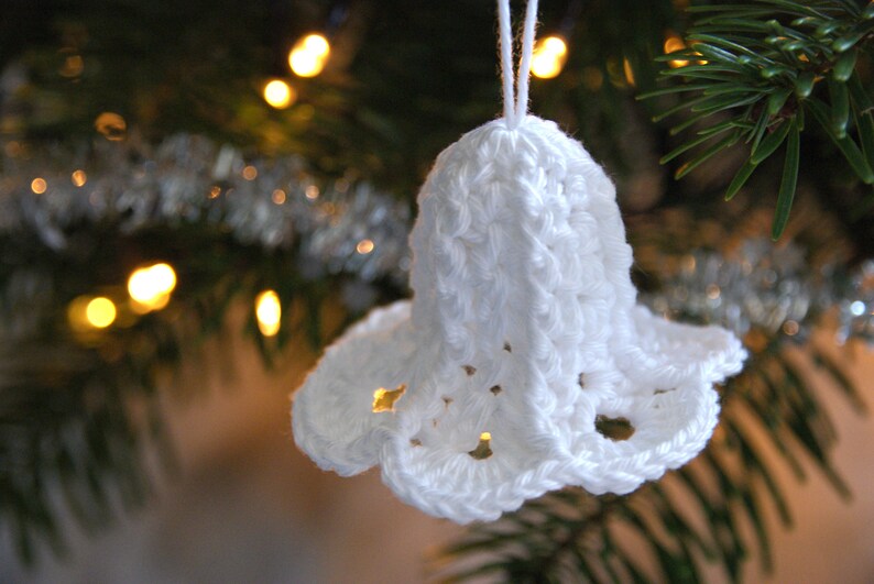 Puede incluir: Adorno de campana de crochet blanco colgado de un &aacute;rbol de Navidad. La campana tiene un dise&ntilde;o delicado y de encaje y es una decoraci&oacute;n navide&ntilde;a tradicional.
